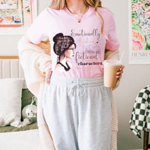 May include: A pink t-shirt with a graphic of a woman with books on her head and the text "Emotionally attached to fictional characters".