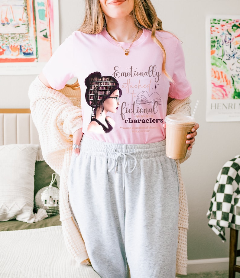 May include: A pink t-shirt with a graphic of a woman with books on her head and the text "Emotionally attached to fictional characters".