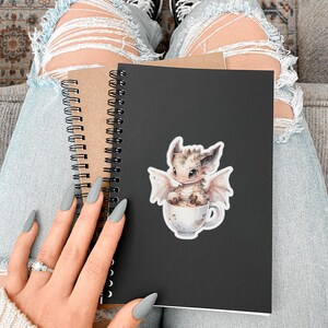 May include: A black spiral bound notebook with a sticker of a small, light brown dragon sitting in a white mug with a brown liquid. The dragon has light pink wings and a light brown body with dark brown spots. The notebook is held by a person wearing blue jeans and a white sweater.