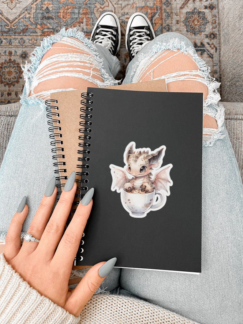 May include: A black spiral bound notebook with a sticker of a small, light brown dragon sitting in a white mug with a brown liquid. The dragon has light pink wings and a light brown body with dark brown spots. The notebook is held by a person wearing blue jeans and a white sweater.