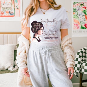 May include: White t-shirt with a graphic of a woman with a stack of books on her head and the text "Emotionally attached to fictional characters".