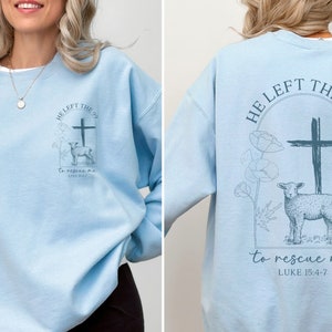 Pu&ograve; includere: Felpa blu chiaro con una stampa grafica di una croce e un agnello. Il testo sulla felpa recita "He left the 99 to rescue me Luke 15:4-7".
