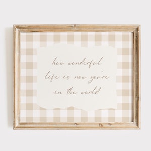 May include: A framed print with a beige and white gingham pattern background. The print features the text "how wonderful life is now you're in the world!" in a brown cursive font.