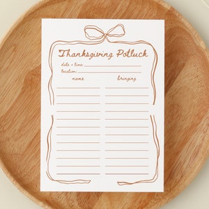 May include: A white Thanksgiving Potluck invitation card with brown text and a decorative border. The card includes spaces for date, time, location, name, and what guests are bringing. The card is displayed on a wooden plate.