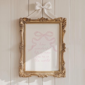May include: A gold framed print with a pink watercolor bow and the text "how wonderful life is now you're in the world".