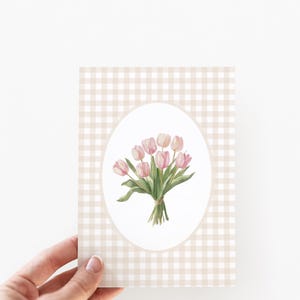 Pu&ograve; includere: Una carta con uno sfondo a quadri vichy e un ovale bianco al centro. L'ovale contiene un'illustrazione ad acquerello di un mazzo di tulipani rosa.