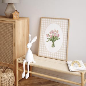 Pu&ograve; includere: Una stampa incorniciata di un mazzo di tulipani rosa con foglie verdi su uno sfondo a quadri vichy beige e bianco. La stampa &egrave; in una cornice di legno ed &egrave; appesa a un muro bianco.
