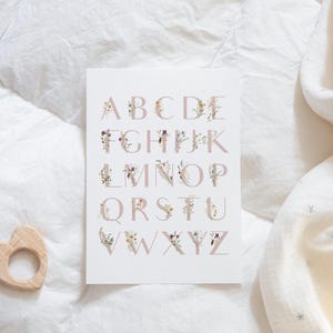 May include: A white card with the alphabet printed in pink with floral designs. The letters are in uppercase and are arranged in alphabetical order. The card is on a white background.