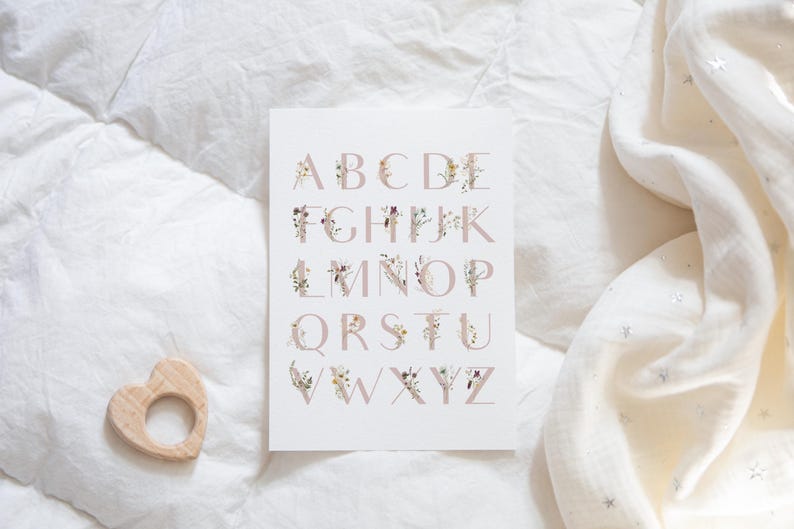 May include: A white card with the alphabet printed in pink with floral designs. The letters are in uppercase and are arranged in alphabetical order. The card is on a white background.