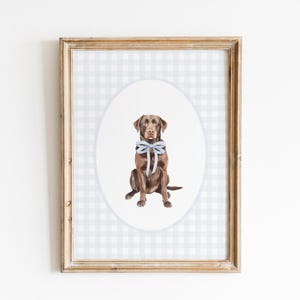 May include: A framed watercolour painting of a brown Labrador Retriever wearing a light blue bow. The painting is on a white and grey gingham background.