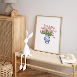 May include: A white ceramic rabbit figurine with long legs sits on a light brown wooden bench. A framed print of pink tulips in a blue and white vase hangs on a white wall behind the bench.