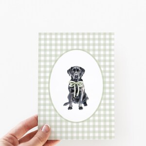 May include: A black Labrador Retriever wearing a light green bow tie sits in a white oval frame against a green and white gingham background.