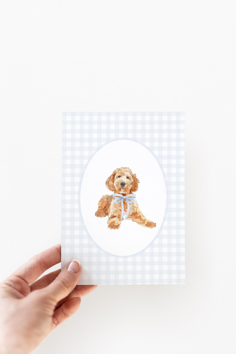 May include: A greeting card with a watercolour illustration of a golden doodle dog wearing a blue bow. The dog is within a white oval on a light blue and white gingham pattern background.