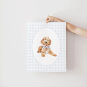 Pode incluir: Impress&atilde;o em aquarela de um c&atilde;o golden doodle com um la&ccedil;o azul, inserida em um oval sobre um fundo xadrez azul claro e branco. A obra de arte &eacute; segurada por uma m&atilde;o.