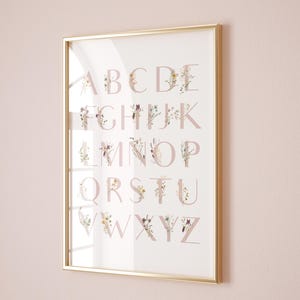 May include: A gold framed print with a white background and a pink alphabet with floral designs. The letters are A through Z.