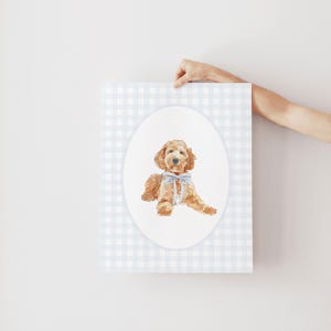 May include: A watercolour illustration of a golden doodle dog wearing a blue bow, set within an oval frame. The background is a light blue and white gingham pattern. The artwork is held up by a person's hand.