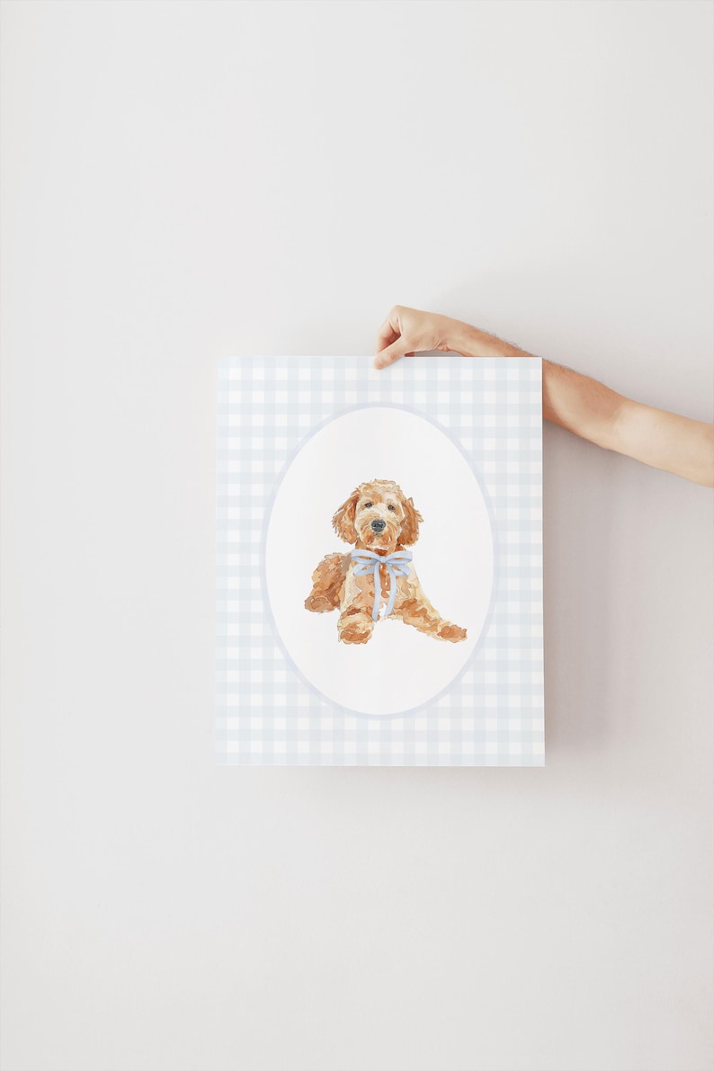 May include: A watercolour illustration of a golden doodle dog wearing a blue bow, set within an oval frame. The background is a light blue and white gingham pattern. The artwork is held up by a person's hand.