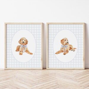 May include: Two framed prints with watercolour illustrations of a golden doodle dog. Each dog is lying down with a light blue bow. The background is a light blue and white checkered pattern.