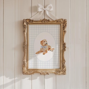 May include: A framed watercolour print of a golden doodle puppy wearing a blue bow. The print is set against a light blue and white gingham background within an ornate gold frame. A white bow and ribbon are attached to the top of the frame for hanging.