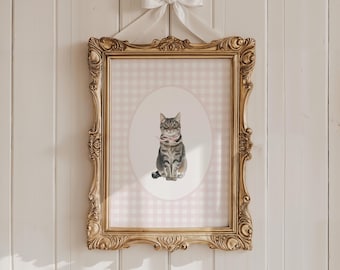 May include: A framed cat art print. The print features a watercolor painting of a gray tabby cat with a pink scarf, set against a pink and white gingham background. The ornate gold frame is suspended by a white bow.