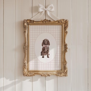 May include: A framed dog portrait with a gold ornate frame and a white bow. The artwork features a brown dog with a pink bow, set against a pink and white gingham background within an oval. The frame is hanging on a white wooden wall.
