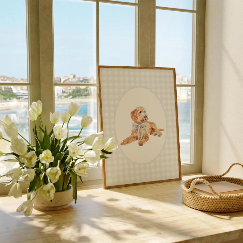 Pode incluir: Uma impress&atilde;o em aquarela emoldurada com um c&atilde;o golden doodle com um la&ccedil;o azul. A obra de arte est&aacute; sobre um padr&atilde;o de xadrez vichy bege claro e emoldurada em madeira natural. Um buqu&ecirc; de tulipas brancas est&aacute; perto de uma janela.