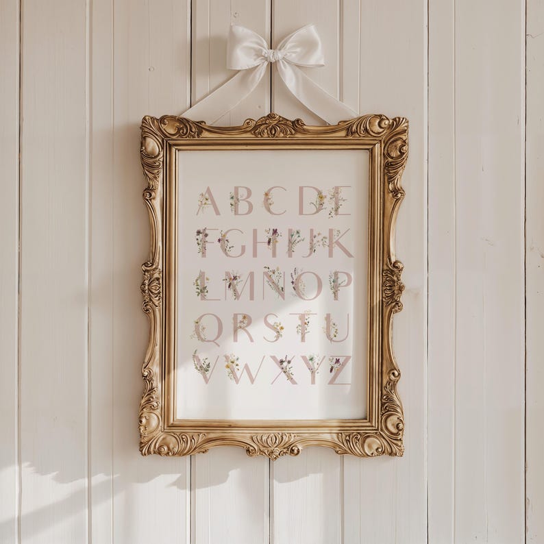 May include: A gold framed print with a white background and a floral alphabet design. The letters are in a light pink color and are surrounded by small floral illustrations. The letters are arranged in alphabetical order from A to Z.