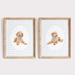May include: Two framed prints. Each features a watercolour illustration of a golden-coloured dog wearing a blue bow. The dog is set against an oval on a light blue and white gingham background. The frames are light brown.