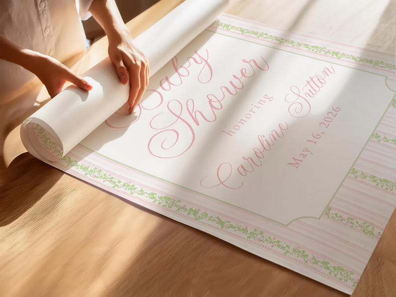 May include: A rolled baby shower banner with pink script text reading "Baby Shower honoring Caroline Sutton May 16, 2026." The banner has a pink and white striped border with green floral accents.