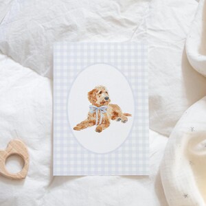 Pode incluir: Ilustra&ccedil;&atilde;o em aquarela de um filhote de golden doodle com um la&ccedil;o azul, emoldurado em um oval sobre um fundo xadrez vichy azul claro. Um mordedor de madeira em forma de cora&ccedil;&atilde;o est&aacute; no canto inferior esquerdo.