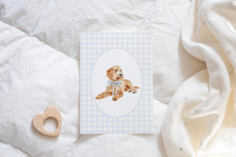 Pode incluir: Ilustra&ccedil;&atilde;o em aquarela de um filhote de golden doodle com um la&ccedil;o azul, emoldurado em um oval sobre um fundo xadrez vichy azul claro. Um mordedor de madeira em forma de cora&ccedil;&atilde;o est&aacute; no canto inferior esquerdo.