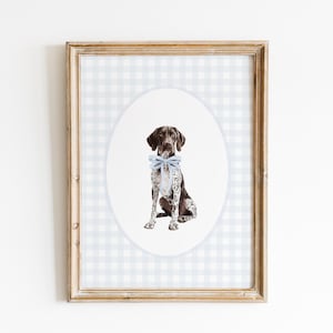 May include: A framed print of a German Shorthaired Pointer, seated and wearing a light blue bow tie. The dog is brown and white, set against a white oval and a light blue gingham background. The frame is light brown.