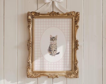May include: A rectangular wall art piece featuring a portrait of a tabby cat centered within an oval on a light pink and white gingham background. The print is housed in an ornate gold frame and suspended by a white satin ribbon tied in a bow.