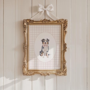 May include: A framed watercolour print of a dog wearing a pink bow tie, set against a pink and white gingham background. The ornate gold frame is suspended by a white satin bow. The artwork is mounted on a white wooden wall.