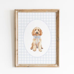 May include: A framed watercolor print of a golden-colored dog with a blue bow tie. The dog is centered on a white oval against a light blue and white gingham pattern. The frame is a light brown color.
