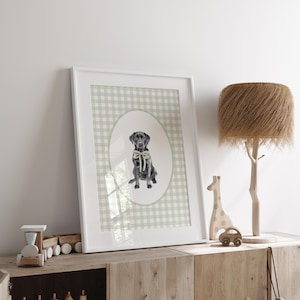 May include: A framed print of a black Labrador Retriever wearing a green bow tie, sitting in a green and white gingham pattern oval. The print is in a white frame and is hanging on a white wall.
