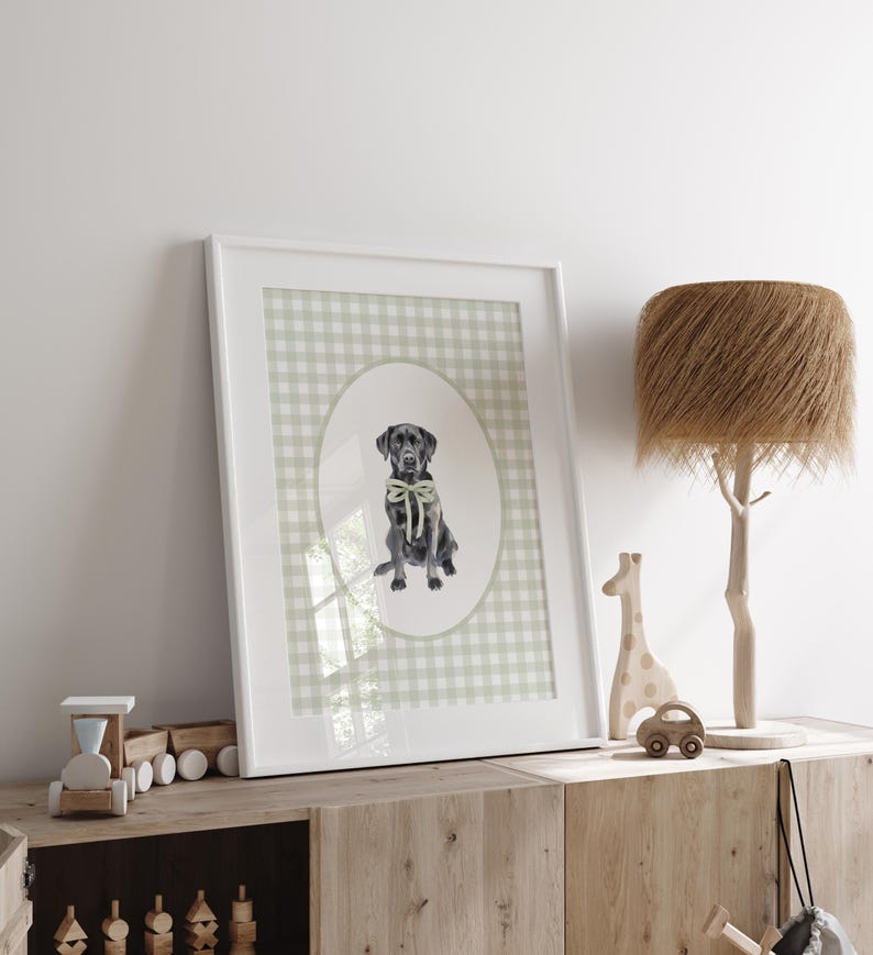 May include: A framed print of a black Labrador Retriever wearing a green bow tie, sitting in a green and white gingham pattern oval. The print is in a white frame and is hanging on a white wall.