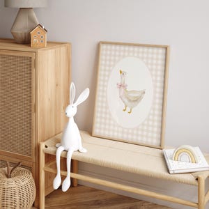 May include: A white goose with a pink bow sits in an oval frame on a beige and white gingham background. The print is framed in a light wood frame.