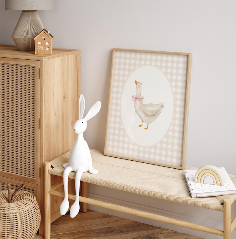 May include: A white goose with a pink bow sits in an oval frame on a beige and white gingham background. The print is framed in a light wood frame.