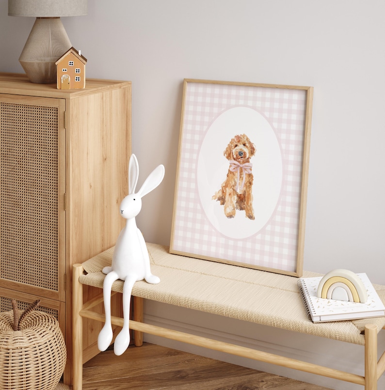 May include: A watercolor painting of a golden doodle dog wearing a pink bow, framed in a pink and white gingham print. The painting is hanging on a white wall above a wooden bench.