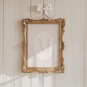 May include: A gold ornate frame with a pink and white gingham pattern and a large white oval with the letter "M" in pink inside. The frame is hanging on a white wood wall.