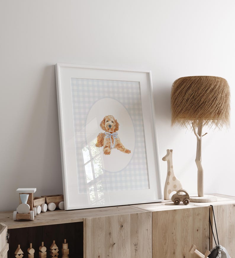 May include: A framed watercolour print featuring a golden doodle dog with a blue bow. The artwork is set against a grey and white gingham background within a white frame. The print is displayed on a wooden cabinet with wooden toys and a tree-shaped lamp.