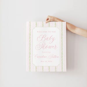 May include: A baby shower sign with a pink and white striped pattern and green floral borders. The sign displays the text "Welcome to the Baby Shower honoring Caroline Sutton May 16, 2026" in a decorative font.
