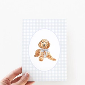 Pode incluir: Ilustra&ccedil;&atilde;o em aquarela de um c&atilde;o golden doodle com um la&ccedil;o azul claro, emoldurada em um oval. O fundo apresenta um padr&atilde;o xadrez vichy azul claro e branco. A imagem est&aacute; em um cart&atilde;o.