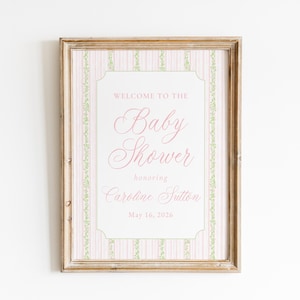 May include: A framed baby shower announcement. The sign has a pink and white striped background with a decorative border. The text reads "Welcome to the Baby Shower honoring Caroline Sutton May 16, 2026."