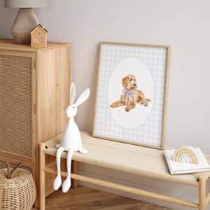 Pode incluir: Uma impress&atilde;o emoldurada em aquarela de um c&atilde;o dourado com uma fita azul, em um fundo xadrez vichy azul claro e branco. A obra de arte &eacute; exibida em uma moldura de madeira clara. A imagem faz parte de um tema de decora&ccedil;&atilde;o de ber&ccedil;&aacute;rio.