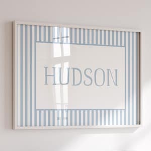 May include: Framed wall art featuring the name "HUDSON" in light blue, centered on a white background. The frame has a light blue and white striped pattern. The artwork is encased in a white frame.