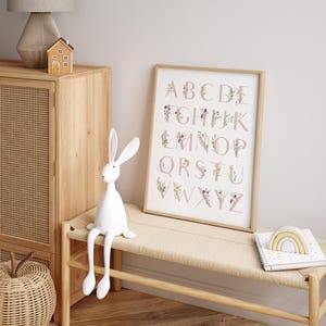 May include: A framed alphabet print with pink letters and floral designs. The letters are surrounded by delicate flowers and greenery. The print is hanging on a white wall above a wooden bench with a white rabbit figurine sitting on it.