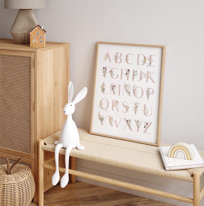 May include: A framed alphabet print with pink letters and floral designs. The letters are surrounded by delicate flowers and greenery. The print is hanging on a white wall above a wooden bench with a white rabbit figurine sitting on it.