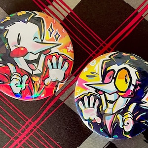 May include: Two circular, colourful badges featuring a cartoon character with a white face, red nose, and yellow glasses. The character is wearing a red suit and has a wide smile. The badges have a holographic effect.